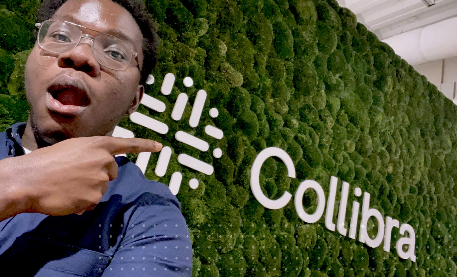 Collibra intern Sam Mensah points his finger at living wall with a large Collibra logo.