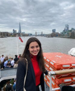 Dori Kovacs stands smiling on a boat floating on the River Thames. London's Tower Bridge is in the distance behind her.