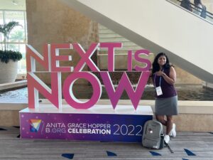 Vidhya stands in front of a sign that says "next is now" at the Grace Hopper Celebration