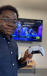 Sam Mensah holds a video game controller in front of a TV screen that displays the video game FIFA22.